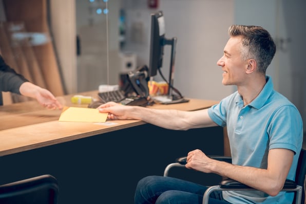 An employee hands a receipt to a man in a wheelchair, demonstrating the importance of ADA compliance when designing ballistic barriers.